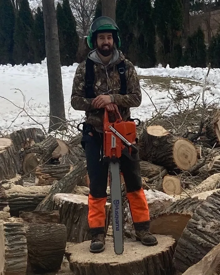 Tyler Smith, owner of Rooted, standing on a fresh stump with his chainsaw, cut rounds piled around him